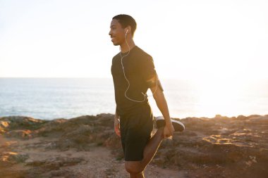 Bin yıllık neşeli Afro-Amerikan erkek telefon ve kulaklıkları ile bacak esnetme egzersizleri yapıyor, gündoğumunda okyanus sahilinde müzik dinliyor, profil çıkarıyor. Uygulama, sabahları egzersiz, vücut bakımı.