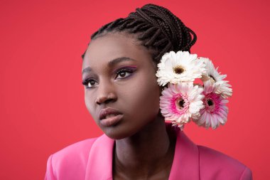 Artistik makyajlı genç zenci kadın portresi ve kırmızı stüdyo arka planında gerbera pozu, kafasında çiçekler ve burnunda piercing olan Afrikalı Amerikalı kadın modelin moda çekimi.