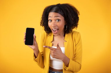 Duygusal Afrikalı Amerikalı kadın elinde boş ekranla akıllı telefonu işaret ediyor, sarı stüdyo arka planında izole edilmiş poz veriyor, güzel bir online teklif sunuyor,