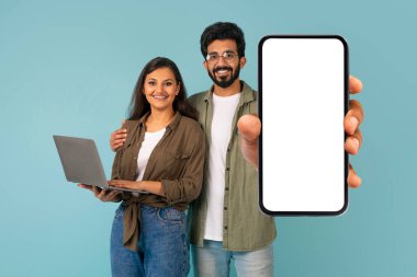 Mavi arka planda gelişigüzel poz veren, modern bilgisayar kullanan, beyaz ekran, maket, kopyalama alanı olan büyük telefonları gösteren neşeli, neşeli milenyum melez bir çift.