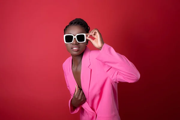 stock image Fashion Shot Of Young Black Woman Wearing Pink Jacket And Stylish Sunglasses Standing Over Red Background, Gramorous African American Female Model Looking At Camera While Posing In Studio, Copy Space