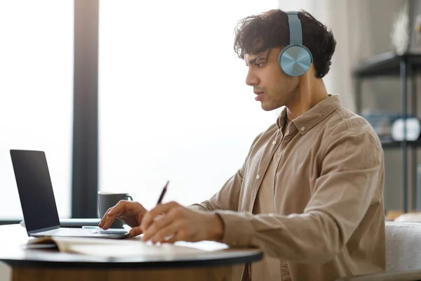 ノートパソコンを使った本格的なアラビア人男性オンラインで働くとヘッドフォンを着用ノートを取り 現代のオフィスでデスクに座っています 学生の男のサイドビューショット遠隔屋内学習を書き込みます Learning — ストック写真