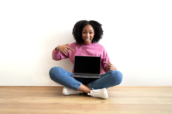 木製の床に座っているクレジットカードで若いアフリカ系アメリカ人の巻き毛の女性を笑顔 リビングルームのインテリアのラップトップで指を指しています 推薦オンラインブログ ウェブサイト 自宅での仕事のためのデバイス — ストック写真