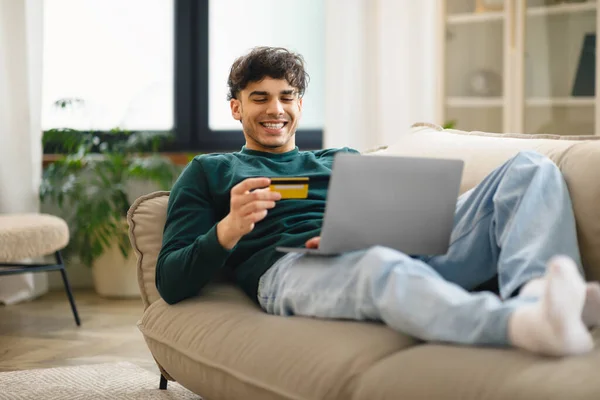 网上购物优惠 快乐的中东年轻人持信用卡在家用笔记本电脑躺在沙发上付款 伟大的销售和电子商务概念 选择性焦点 — 图库照片