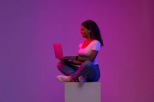 Smiling Black Woman Using Laptop While Sitting On Big Cube In Neon Light, Happy Young African American Female Working Online Or Study With Computer Over Purple Studio Background, Copy Space
