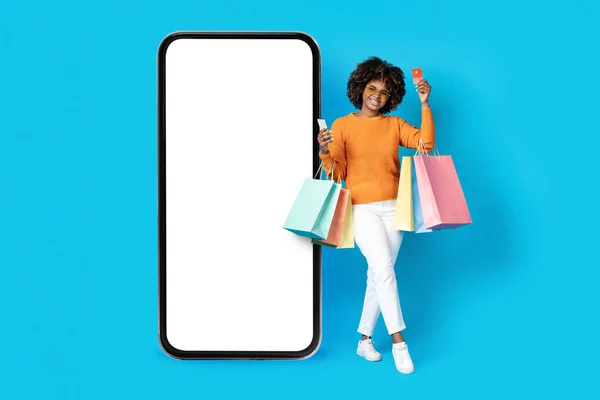 时尚漂亮的年轻黑人女士 穿着休闲装 站在巨大的智能手机旁 白色的空白屏幕边 手里拿着东西 上网购物 用手机和银行卡 蓝色背景 装模作样 — 图库照片