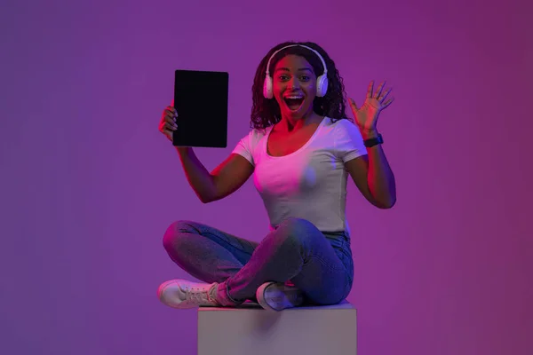 ネオンライトで大きなキューブに座っている間興奮した黒人女性がブランクデジタルタブレットを保持します ワイヤレスヘッドフォンを身に着けている陽気な若いアフリカ系アメリカ人女性は 新しいアプリやウェブサイトをお勧めします モックアップ — ストック写真