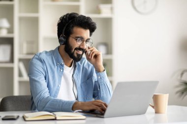 Evde dizüstü bilgisayarla kulaklık takan gülümseyen Hintli genç adam, masada oturan yakışıklı Doğulu adam, bilgisayarda Webinar 'ı izliyor, çevrimiçi eğitimin ve uzaklık öğrenmenin tadını çıkarıyor, boşluğu kopyalıyor.