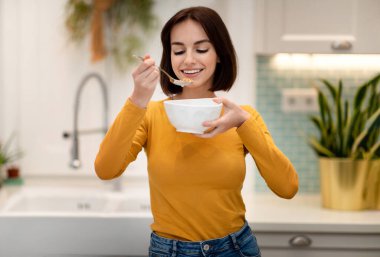 Sağlıklı, neşeli, kısa saçlı, günlük kıyafetli, elinde beyaz kase ve kaşıkla genç bir bayan. Evde kahvaltı yapan, tahıl, yulaf ezmesi ya da mısır gevreği yiyen mutlu bir kadın.