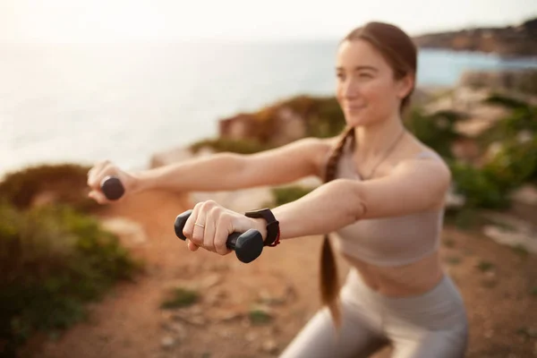 スポーツウェアの肯定的なエネルギーの若い白人女性はダンベルと腕運動をし 海のビーチでのトレーニングを楽しみ やる気を起こさせる ボディケア ヘルスケア フィットネス ワークアウト屋外 — ストック写真