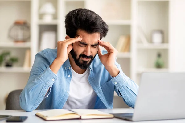 Tired Young Indian Freelancer Guy Having Headache While Working In Home Office, Millennial Eastern Man Sitting At Desk, Rubbing Temples And Frowning, Suffering From Acute Migraine, Copy Space