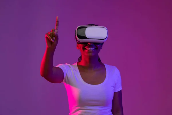 Excited Black Woman Wearing Vr Headset Enjoying Virtual Reality Experience, Smiling African American Female Touching Air With Finger, Standing In Neon Light Over Purple Studio Background, Copy Space