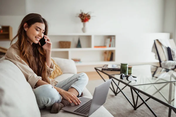 Akıllı telefondan arayan, minimalist oturma odasında bilgisayarda yazan mutlu Avrupalı bir bayan. Serbest meslek, iş ve iş evde, birden fazla iş için aygıt