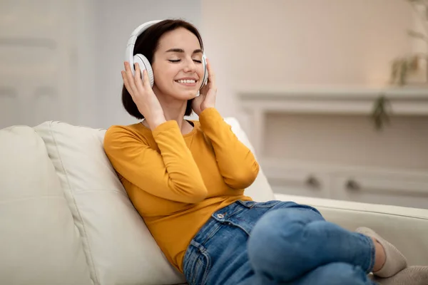 穿着休闲装的无忧无虑的漂亮黑发女子坐在沙发上 家中用白色无线耳机 闭着眼睛听音乐 复制空间 — 图库照片