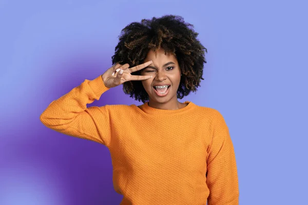 stock image Portrait of cheerful cool stylish young black woman with bushy hair in orange sweater posing on purple studio background, winking and showing peace gesture next to her eye