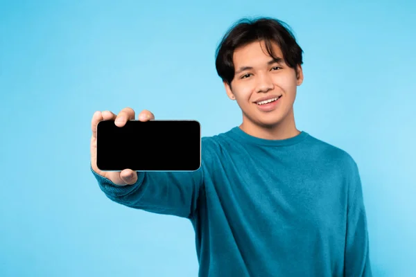 青の背景の上に携帯電話のブランク画面を示す陽気な中国のティーンの男 スマートフォン広告を保持している学生のスタジオショット素晴らしいモバイルオファー 新しいアプリ広告 モックアップ — ストック写真