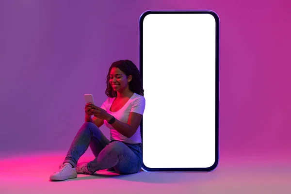 Mobile Offer. Smiling Black Woman Using Smartphone And Leaning At Big Blank Cellphone With White Screen, Happy African American Female Sitting In Neon Light On Purple Background, Mockup