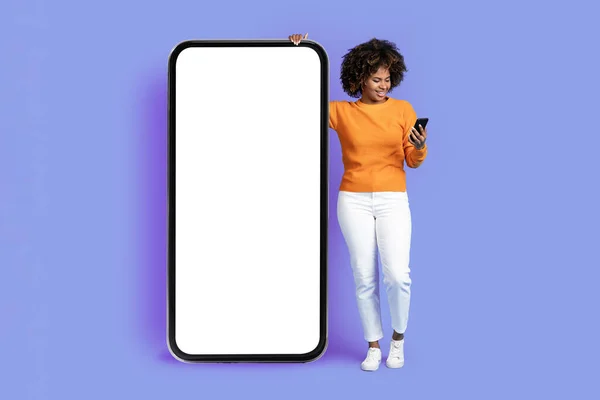 活泼时尚的年轻非洲裔美国女人 一头浓密的头发 站在巨大的智能手机旁边 白色的空白屏幕 使用手机 在紫色的工作室背景上微笑 喜欢漂亮的应用程序 — 图库照片