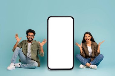Pozitif, sevimli, genç, güzel, doğulu çift güzel kadın ve yakışıklı adam büyük cep telefonunun yanında beyaz ekranlı, el kol hareketi, flört uygulaması için model, mavi arka plan.