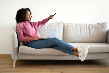Gülümseyen Afro-Amerikan kıvırcık kadın uzaktan kumandayla koltukta oturuyor. Minimalist oturma odasında boş zaman ve televizyon programının tadını çıkarıyor. Rahatla, evde dinlen, film akşamı.