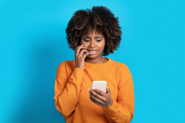 Korkmuş, kıvırcık saçlı, saçları gür, cep telefonu ekranına bakıp yüzünü buruşturan, dudaklarına dokunan güzel, kıvırcık, siyah bir kadın. Korkunç haberler okuyor, mavi stüdyo arka planında izole edilmiş.