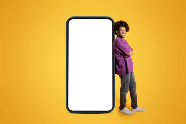 身穿紫色T恤的快乐而自信的非洲裔美国卷曲男子站在巨大的智能手机旁边 屏幕空白 背景为黄色 广告和提供 网站和销售 — 图库照片