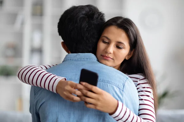 Pretty long-haired young eastern woman chatting with guys on dating app via smartphone while hugging her boyfriend, home interior, copy space. Cheating, deception, dishonesty in relationships