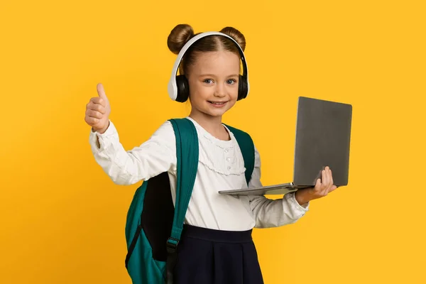 在线教育 可爱的小女孩 带着笔记本电脑和耳机出现在相机前 微笑着享受远程学习 站在黄色工作室的背景下 — 图库照片