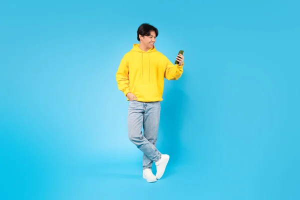 新しいモバイルアプリで携帯電話を使用して韓国の10代の男の子は青の背景に立っている ソーシャルメディアアプリケーションで陽気な男のテキストとスクロールのスタジオショット デジタルレジャーコンセプト — ストック写真