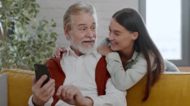 Akıllı telefondan haberleri okuyan mutlu kıdemli bir adam. Neşeli torunu arkadan sarılıp onu kucaklıyor. Evde birlikte vakit geçiriyorlar.