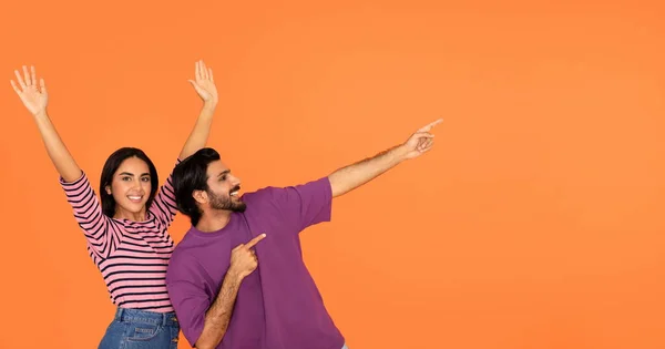 幸せな陽気な感情的な千年紀のインドのカップルが一緒に楽しみを持っているとオレンジのスタジオの背景にテキストや広告のための空のスペースを示す 素晴らしいオファー ウェブバナーを指して — ストック写真