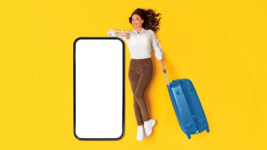 Seyahat Başvurusu İlanı. Sarı Stüdyo Arkaplanı üzerinde mavi bavul taşıyan boş ekran pozuyla büyük telefona yaslanan mutlu gezgin kadın. Turizm ve Gadget Konsepti. Panorama, Mockup