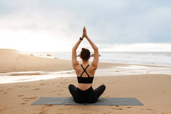 户外瑜伽 女人双手抱头沉思 坐在沙滩上的健身垫上 练习近海静坐 回头看 — 图库照片