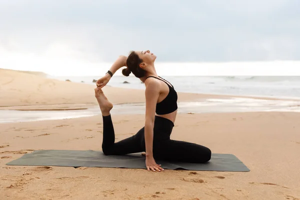 年轻女子在海边的健身垫上伸展和做瑜伽 穿着黑色运动服 喜欢户外晨练 — 图库照片