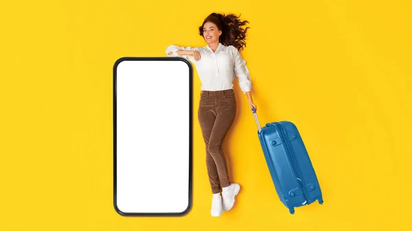 旅行アプリケーション広告 黄色のスタジオの背景に青いスーツケースを保持ポーズを空の画面で大きな携帯電話でリースハッピー旅行者の女性 観光とガジェットの概念 パノラマ モックアップ — ストック写真