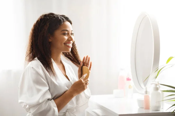 Black Smiling Female Combing Her Beautiful Curly Hair With Bamboo Brush At Home, Attractive African American Woman Sitting At Dressing Table And Looking In Mirror, Making Daily Beauty Routine
