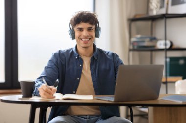 Laptop kullanan, kameraya gülümseyen, modern ofiste masada oturan kulaklık takan neşeli Arap adam. Başarılı Öğrenci Öğrenme İnternetinin Portresi. E- Öğrenme