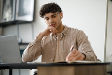 E-öğrenme. Orta Doğulu adam internetten öğreniyor dizüstü bilgisayarında oturmuş kulaklık takıyor. İçerideki masada uzaktan kumandayla poz veren bir adam. Uzaklık Eğitimi