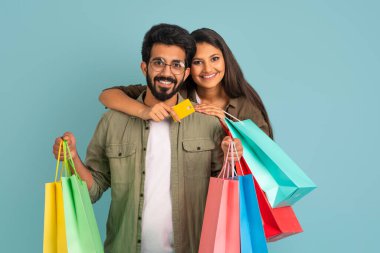 Sevimli, neşeli, genç Hintli çift mutlu erkek ve kadın birlikte alışveriş yapıyor, renkli alışverişler yapıyorlar. Mavi stüdyo arka planında izole edilmiş bir banka kartı. E-ticaret kavramı