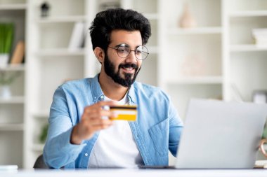 Evde dizüstü bilgisayar ve kredi kartı kullanarak gülümseyen Hintli erkek, iş yerinde otururken internetten ödeme yapan ya da internetten alışveriş yapan yakışıklı Doğulu serbest yazar,