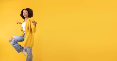 Eğleniyorum. Mutlu genç siyahi bayan zıplıyor, dans ediyor ve fotokopi odasına bakıyor, iyi bir ruh hali içinde, sarı arka planda izole edilmiş, stüdyo çekimi, panorama