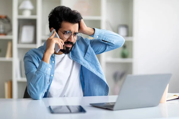 紧张的印度男性自由职业者在家里工作时对电脑有问题 灰心丧气的东方人在客户的支持下一边用手机聊天 一边用震惊的目光看着笔记本电脑屏幕 — 图库照片