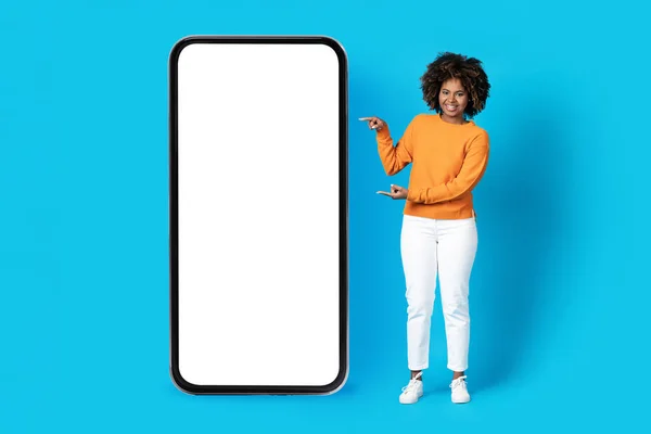 快乐快乐的年轻黑人女人 浓密的头发 拿着大手机 白色的空屏幕 指着小玩意 对着蓝色的工作室背景微笑 推荐漂亮的应用程序 — 图库照片