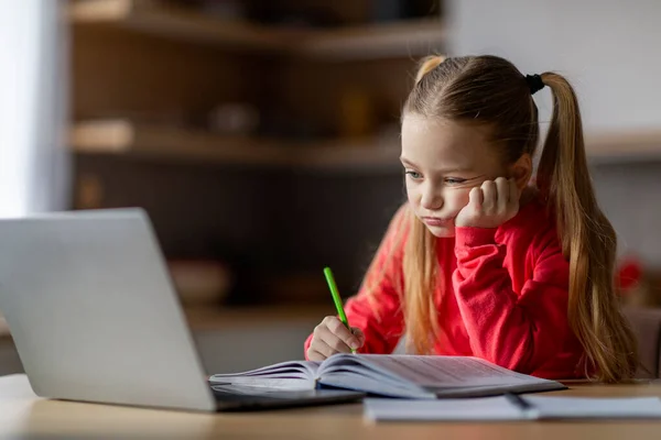 stock image Portrait of bored little girl doing homework with laptop and book, tired preteen female child holding pencil and looking at computer screen, preparing for exam test, having problems with education