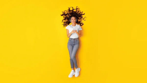 音楽アプリケーション スタジオで黄色の背景の上にイヤフォンを身に着けている音楽をオンラインポージングに電話を保持興奮した女性 歌を楽しむリラックスしている女性 パノラマ — ストック写真