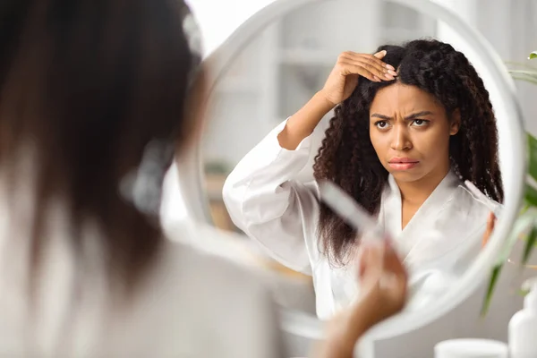Upset Black Female Looking At Mirror At Home And Touching Her Hair Roots, Frustrated Young African American Woman Suffering Dandruff Or Hairloss Problem, Selective Focus On Reflection