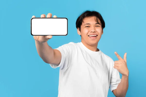 日本の10代の若者が携帯電話を空画面で表示し 青いスタジオの背景に指の広告アプリケーションを指して幸せ 大規模なモバイルオファー広告 モックアップ — ストック写真
