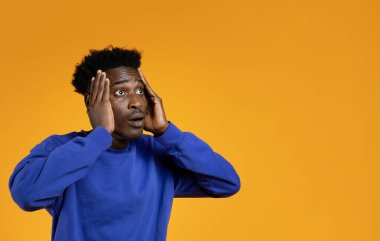 Şok olmuş, mavi kazaklı yakışıklı genç adam reklam için fotokopi odasına bakıyor, yüzüne dokunuyor, sarı stüdyo arka planında şaşkınlığı gösteriyor, web-afişi