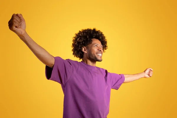 Smiling excited adult african american curly man in purple t-shirt raises arms to sides enjoys freedom on yellow studio background. Great news, victory emotions, ad and offer