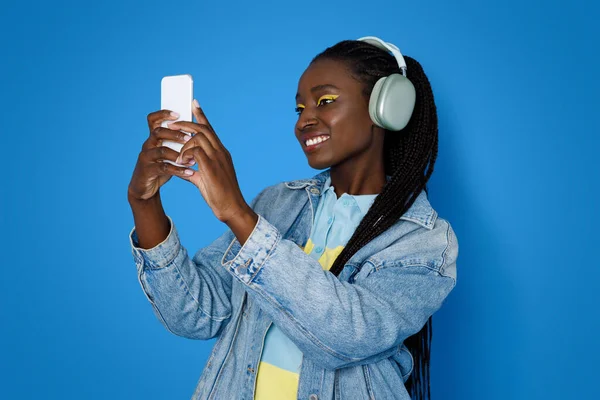 电信应用程序 穿着休闲装的年轻时髦黑人女人 使用现代无线立体声耳机和智能手机 和朋友们在网上聚会 蓝色工作室背景 — 图库照片